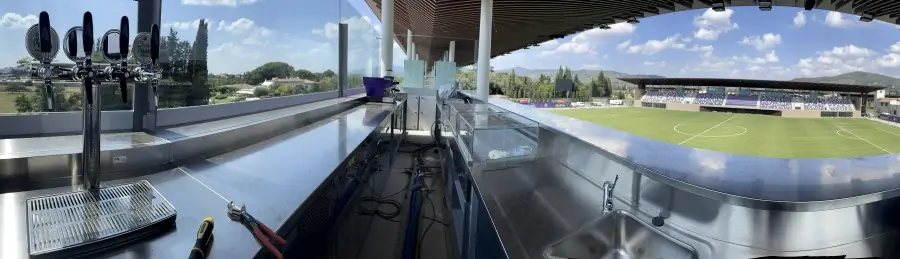 Panoramica dal bancone del bar del Viola Park: la colonna di rubinetti in primo piano, dietro le vetrate il campo e la tribuna.